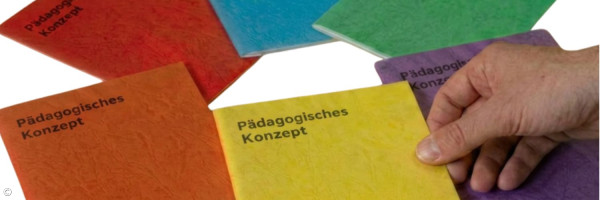 Das Bild zeigt mehrere farbige Broschüren mit dem Titel „Pädagogisches Konzept“, die fächerförmig auf einer hellen Fläche ausgebreitet sind. Die Hefte sind in verschiedenen Farben gehalten, darunter Rot, Blau, Grün, Gelb, Orange und Violett. Im Vordergrund greift eine Hand nach einer gelben Broschüre. In der rechten unteren Ecke ist ein kleines Logo zu sehen.